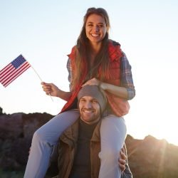 Portrait of young tourist couple celebrating American freedom: beautiful girl in  hiking outwear sitting on her boyfriends shoulders smiling cheerfully to camera and waving small American flag at sunset